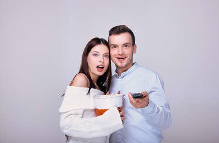 A young girl in a white sweater opened her mouth in surprise, holds a bucket of popcorn in her hands and snuggles up to a cheerful guy in a blue shirt who is holding a TV remote control.の写真素材