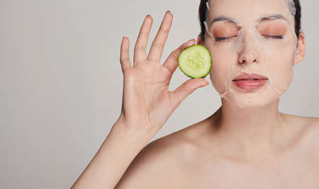 Close up calm sophisticated beauty girl in a moisturizing mask with a fresh cucumber on the face in the background serious with close eyes, hands holds a cucumbers in both hands near her faceの写真素材