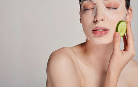 Middle frame photo, gentle sophisticated calm girl in a moisturizing mask with a fresh cucumber on the face in the background serious with closed eyes and one left hand near her face touching cucumberの写真素材