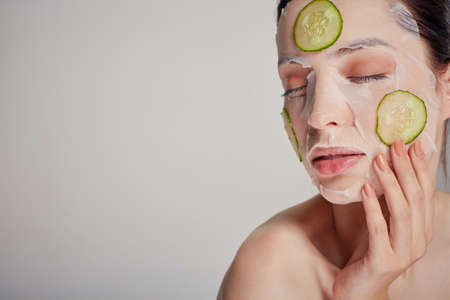 Close up gentle sophisticated girl in a moisturizing mask with a fresh cucumber on the face in the background serious with closed eyes, hand near her faceの写真素材
