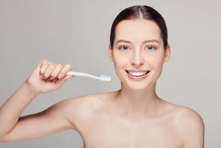 Young pretty girl looking straight and smiling with a toothbrush in her hands nearly her face with white teeth, with brown hair with clean fresh skin posing on a gray studio background, copy spaceの写真素材