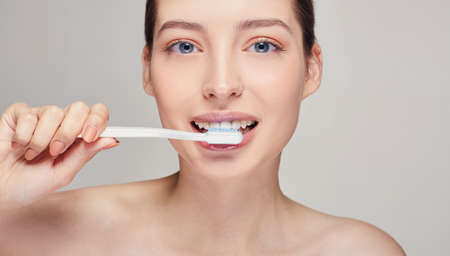 Looking straight, nude make up, copy space. Young pretty girl with a toothbrush in her hands nearly her mouth with white teeth, with brown hair with clean fresh skin posing on a gray studio backgroundの写真素材