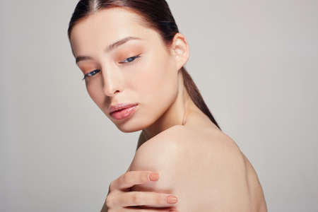 Copy space. Close up. Young beautiful sophisticated girl with blue eyes and full lips holds a hand on his shoulder. Grey background. Dark hair.の写真素材