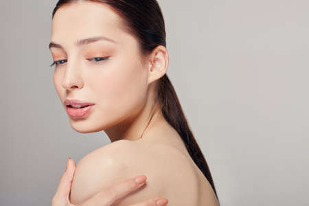 Looking down. Young beautiful pretty girl with blue eyes and full lips holds a hand on his shoulder. Grey background. Dark hair. Copy space.の写真素材