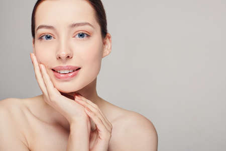 An elegant sophisticated beautiful girl with full lips, dark hair and radiant clean delicate skin on gray background. Lady holding her hands near her face. Looking straight. Skin care. Smiling. Spa.の写真素材