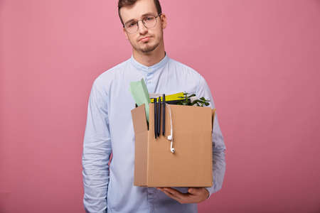 Focus on box. A young pretty guy in a sky blue shirt and computer glasses stands on a pink background with a cardboard box with different things in his hands. Dismissal. Fired.の写真素材