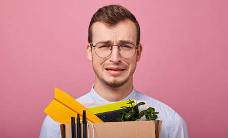 A young pretty guy in a blue shirt and computer glasses stands on a pink background with a cardboard box with pens, plant and different things in his hands. Dismissal. Fired. Man is upset.の写真素材