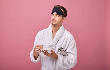 Un sleepy man in a sleeping mask on his head in a white coat with a newspaper under his arm, smiling at the camera with a cup of coffee in hands. Awakening, morning mail. Copy space. Looking sideway.の写真素材