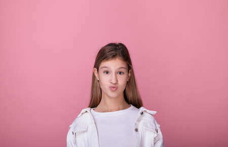 Funny young beautiful girl with long blonde hair in white fashionable clothes folded her lips in a kiss. Teenager girl posing with bright cheerful emotions on a pink background.の写真素材