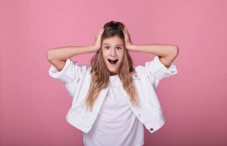 Portrait of a teenage girl, holding her head with two hands, screaming with joy, opened her mouth in surprise. Funny beautiful young blonde posing on a pink background. The emotions of a teenager.の写真素材