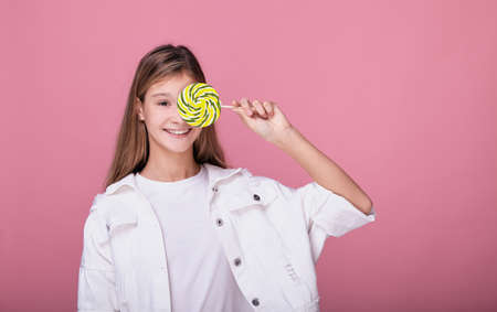 A cute teenager girl in fashionable youth clothes holds a large multi-colored lollipop in her hands and closes her eyes with candy. Young blonde with a candy on a stick posing on a pink background.の写真素材