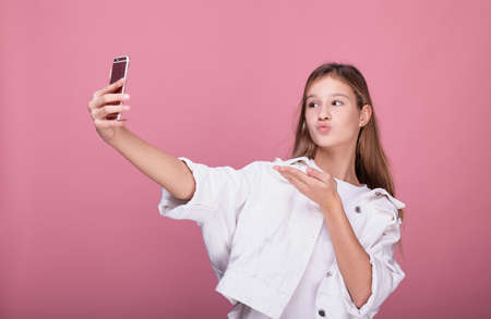 Cute teen girl with long hair in fashionable clothes holds a pink smartphone in her hands and blows a kiss. Girl with a pink smartphone. Children and technology concept.の写真素材