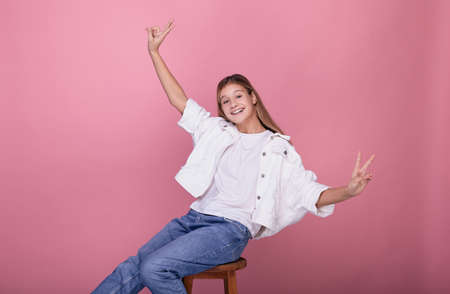 A young smiling girl in blue jeans and a white sweater is sitting on a chair and is showing the peace sign with both hands. A teenage girl smiles, looks straight and shows a peace sign.の写真素材