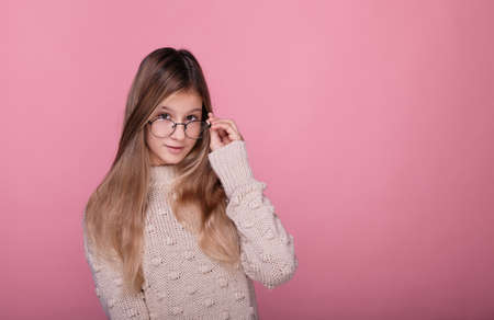 Beautiful stylish teenager girl with long hair in a beige sweater and round glasses looks straight. The girl holds on to the arm of the glasses and playfully looks straight. Cute young schoolgirl.の写真素材