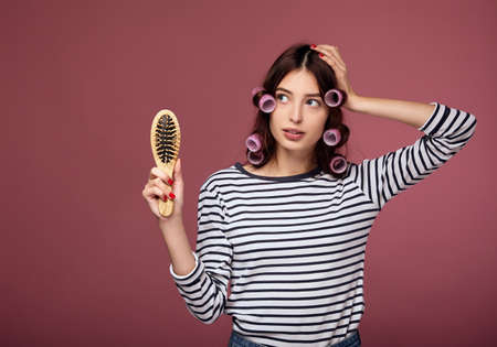 A pensive pretty lady in a striped sweater in curlers stands on a pink background. The lady in the curlers holds a wooden comb in her hand, looks to the side. Hair care, vitamin deficiency, hair loss.の写真素材