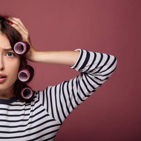 Close-up. A young brunette with blue eyes in a striped sweater and pink curlers clings to her head and is sad. Nice girl in pink curlers. The concept of hair care, healthy hair, fashionable hairstyle.の写真素材