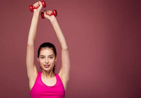 Energetic beautiful white girl with dark hair with blue eyes in a sports pink top and black leggings does fitness with dumbbells. Cute sportswoman goes in for sports with red dumbbells. Copy space.の写真素材