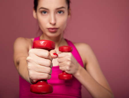 Sporty fit serious brunette with blue eyes in a pink top does exercises with red dumbbells. Beautiful lady is engaged in fitness on a pink background. Concept woman and sport and beauty of the body.の写真素材