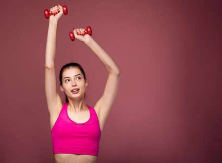 A white slender girl with dark hair and blue eyes in a sports pink top and black leggings is training with red dumbbells. Cute young sportswoman lifted dumbbells up. Copy space.の写真素材