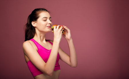 Sensual fitness lady in a pink top and black leggings holds in her hands a large juicy cheeseburger and enjoys biting it. The girl after fitness eats fast food. Copy space.の写真素材