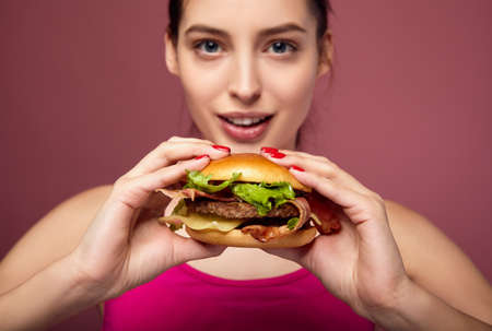 Young pretty athletic lady with blue eyes and fair skin in a pink sports top holds a big juicy burger in her hands. Sports hungry woman with a burger posing on a pink background. Lady on a diet.の写真素材