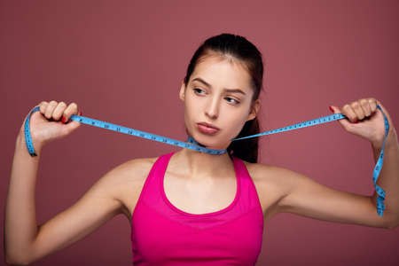 Young athletic brunette with blue eyes and full lips in sportswear is posing against the background with a measuring tape on her neck. A woman measures her parameters. Diet, anorexia, women's health.の写真素材
