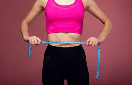 Close up of a young female athletic body in a pink tank top and black leggings. A girl measures her body with a measuring tape. A young lady is using a measuring tape.の写真素材