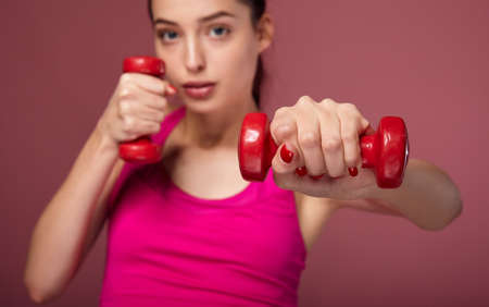 Young slim white girl with blue eyes in a pink top is engaged in fitness with red dumbbells. A beautiful lady holds dumbbells in her hands, stretches one arm forward, looks straight and confidently.の写真素材