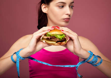 Close-up of a pretty brunette with blue eyes with a blue measuring tape in her hands holds a cheeseburger looking to the side. The girl on a diet eats fast food. The concept of diet eating behavior.の写真素材