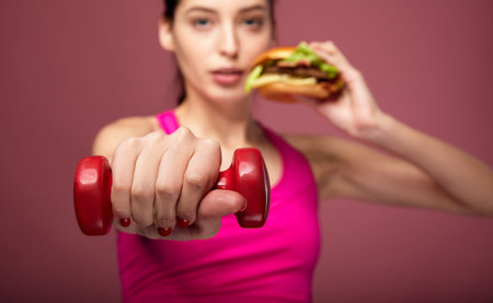 Close up of female slim hand with red dumbbell on pink background. Girl athlete with a burger and...