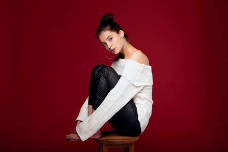 Attractive fashionable glamorous girl with white skin sits on a high wooden chair and looks away. Girl in a fashionable autumn outfit posing on a red background. The concept of fashion and style.の写真素材