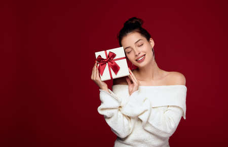 A nice sweet girl with dark hair in a white knitted sweater with bare shoulders bowed her head to the present. Girl with a gift on a red background. Concept Christmas present and Valentine's Day.の写真素材