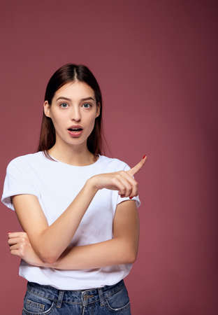 Surprised lovely young female being shocked by interesting unexpected rumor, keeps jaw dropped, indicates at upper right corner, feels stunned, poses against pink background with blank space aside.の写真素材