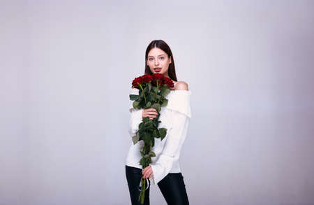 A chic stylish young brown-haired woman with blue eyes in a white knitted sweater and black leather pants holds in his hand a bouquet of long large red roses, looking straight. Copy space.の写真素材