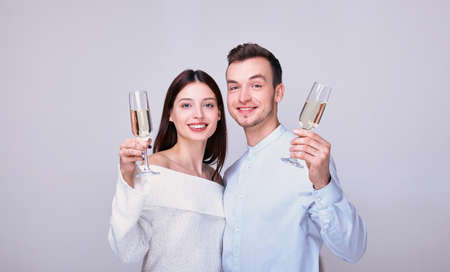 Happy newlyweds drink champagne. A young couple in love in light festive clothes hold glasses of champagne and smile happily, look straight. A couple has fun on white background and drinks champagne.の写真素材