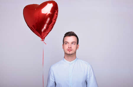 A young man with dark hair in a blue shirt poses against a grey background. A young man holds a large balloon in the shape of a red heart in his hand and looks up. Young in love on Valentine's Day.の写真素材