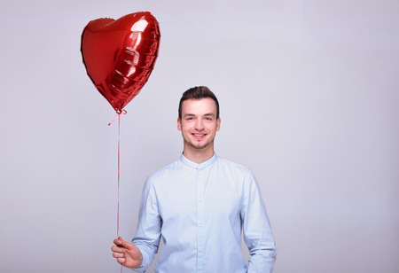 A joyful young white guy with dark hair in a light shirt smiles, looks straight, holds in his hand a large red balloon in the shape of a heart. The concept of Valentine's Day, February 14. Copy space.の写真素材