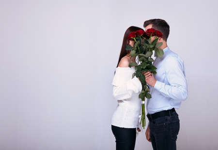 Charming gentle pair of young stylish fashionable lovers kisses, covers his face with a bouquet of red roses. A young happy inspired couple. A romantic date on a white background. Copy space.の写真素材