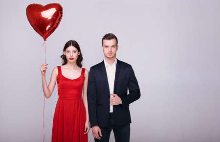 Beautiful romantic couple isolated on a gray background. A serious young woman and a handsome confident man are standing next to heart-shaped balloons in their hands. Happy Valentine's Day.の写真素材