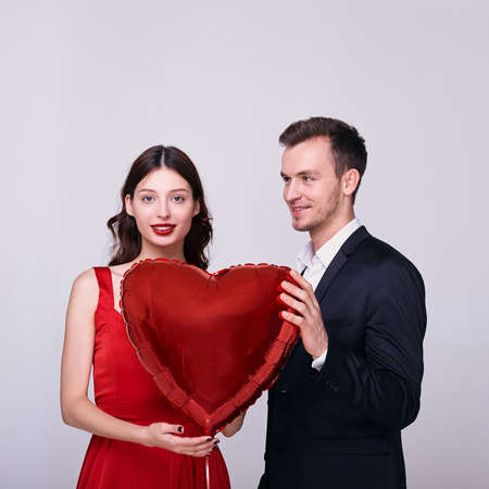 A man in a jacket and a luxurious white woman in elegant evening dresses hold a red bouncy heart and smile. Sexy couple on Valentine's Day on white background. Communication and relationship concept.の写真素材