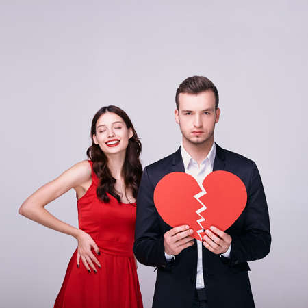 A beautiful bright brown-haired woman with a snow-white smile and red lips hugs Prana in a suit with a red broken heart in her hands. Lovers in quarrel, divorce, broken seven. Love is over.の写真素材