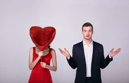 A surprised white man in a suit and white shirt spread his arms to the side, looks straight, stands next to a young white lady in a red dress. A girl holds a red heart balloon instead of a head.の写真素材