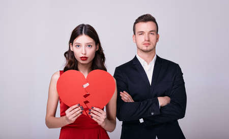 Elegant luxurious pair of young lovers in evening dresses pose with a broken red heart on a white background. The concept of Valentine's Day, broken love, is no longer together.の写真素材