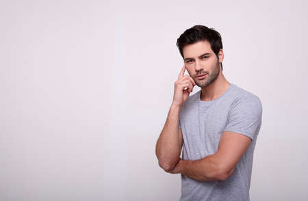 A confident calm white man with a fashionable haircut with dark hair, in a gray T-shirt crossed his arms on his chest. A male model in a grey T-shirt holds his hand near his face and looks straight.の写真素材