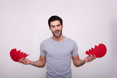 A handsome young bearded guy in a gray T-shirt, sincerely smiling, holds in his hands two halves of a broken heart, standing near a white background. The guy shows a broken love on Valentine's Day.の写真素材