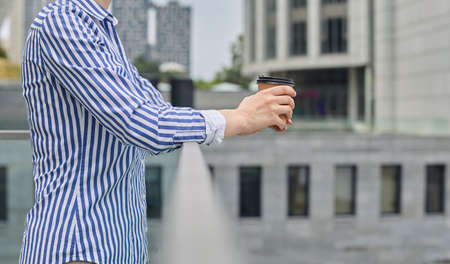 A lady in shirt with a brown glass of takeaway coffee stands in the background of a building in the city, clouding her hands on the railing. Waiting for a meeting, rest, lunch break.の写真素材