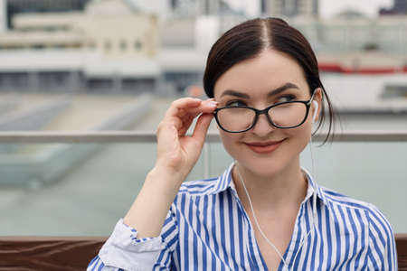 Nice smiling business woman with dark hair tied in a bun in a striped shirt sits in a park on a bench and holds her eyeglasses. Lady looks away and smiles slight.の写真素材