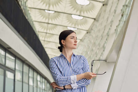 Intelligent serious business lady with dark hair in a blue and white striped shirt with white cuffs on her sleeves and glasses in hand. Girl looks at side against the background of building.の写真素材
