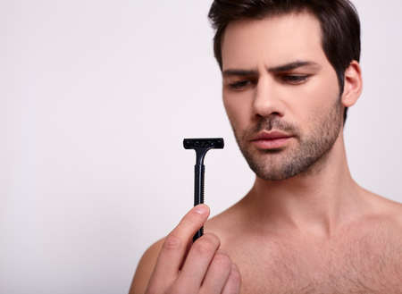 The naked nice brunette guy stands on a white background with a black razor in his hand and looks at her. Taking care of yourself, shaving your face.の写真素材