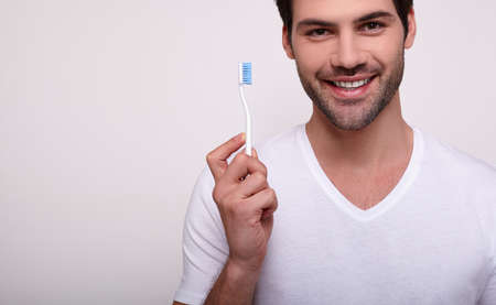 Man with a toothbrush. Cropped image of a young white man in a white T-shirt holding a toothbrush and smiling while standing against a gray background. A man does oral hygiene. Copy space.の写真素材